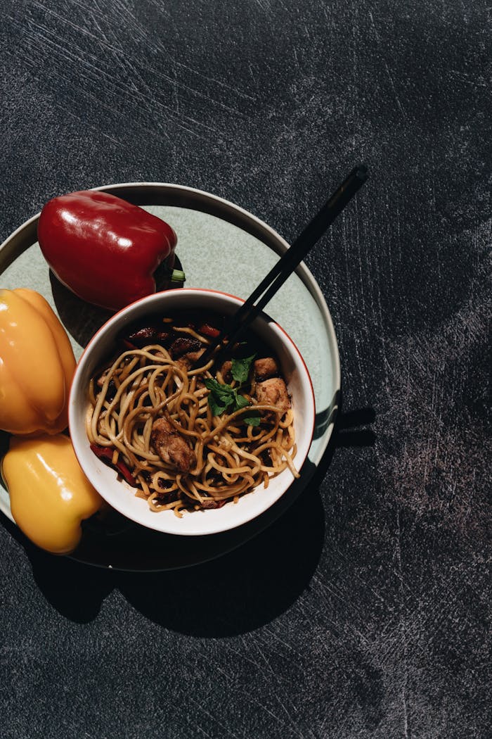 services-01 Top view of a noodle dish with chicken and colorful bell peppers on a dark background.