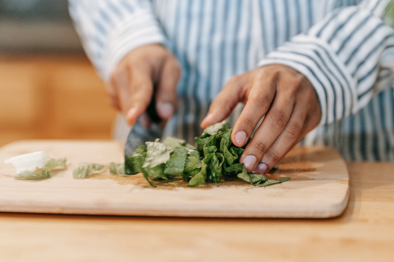 gallery-3 Crop faceless male chef in casual clothes chopping fresh green lettuce leaves with knife on wooden cutting board during cooking process in kitchen